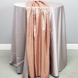 May include: A rose gold sequin table runner draped over a silver table cloth. The runner is made of a soft, shimmery fabric and would add a touch of elegance to any table setting.