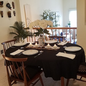 May include: A dining table set for a formal dinner. The round table is covered with a black tablecloth and set with white plates, silverware, and wine glasses. A vase of white flowers and lit candles complete the elegant setting.
