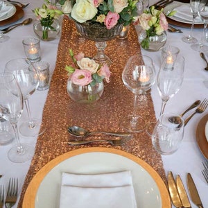 May include: A table setting with a rose gold sequin table runner, white plates, gold chargers, and clear glassware. The centerpiece is a glass vase filled with pink and white roses.