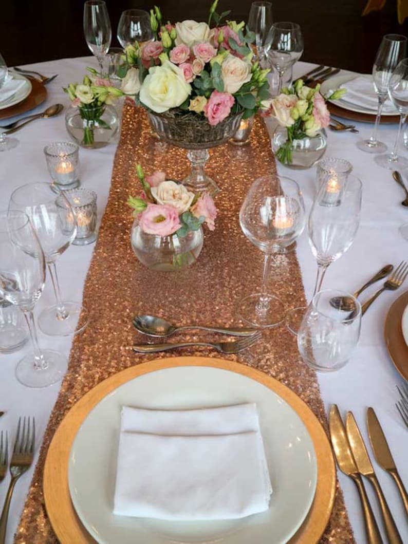 May include: A table setting with a rose gold sequin table runner, white plates, gold chargers, and clear glassware. The centerpiece is a glass vase filled with pink and white roses.