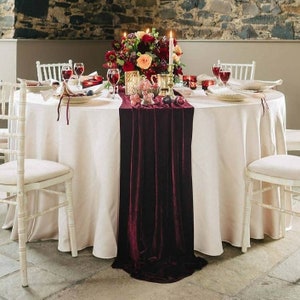A white round tablecloth with a burgundy velvet table runner. The table is set with white plates, wine glasses, and candles. There is a bouquet of red and pink flowers in the center of the table.
