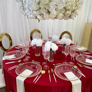 A round velvet red tablecloth with white velvet napkins and gold rimmed glass plates. The table linen fabric is velvet. A large white floral arrangement with white roses and orchids is in the center of the table.