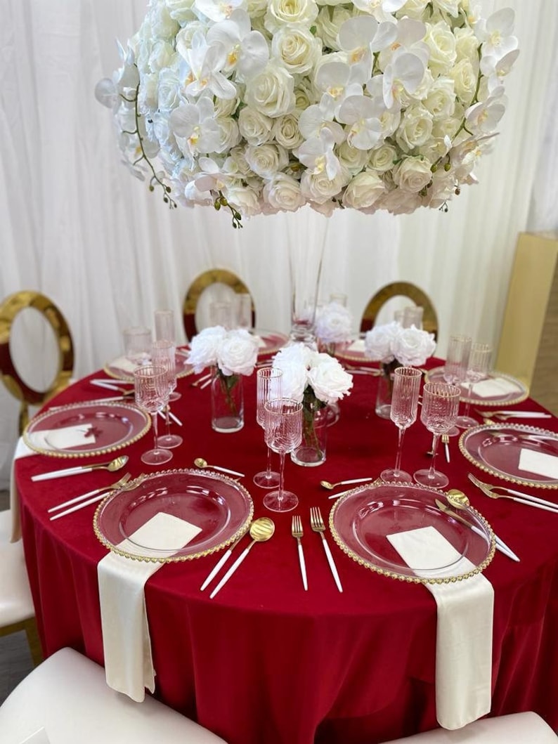 A round velvet red tablecloth with white velvet napkins and gold rimmed glass plates. The table linen fabric is velvet. A large white floral arrangement with white roses and orchids is in the center of the table.