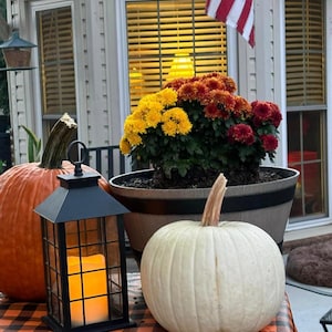 A black and orange plaid fall tablecloth with a orange and a white pumpin on the table, a lantern and orange flowers. The picture is outside of a home that is flying an American flag.