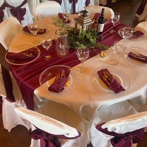 A round table set for a wedding reception with white tablecloths burgundy velvet table runner and napkins. The table is decorated with a centerpiece of greenery and a lantern. There are wine glasses, plates, and silverware set on the table.