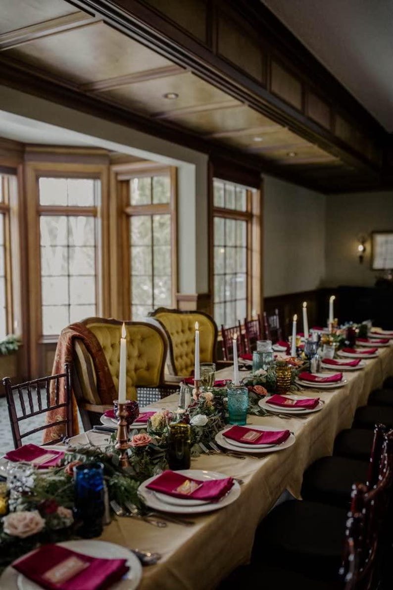 A long wedding reception table with a tan tablecloth and red linen napkins ready for the wedding reception dinner.