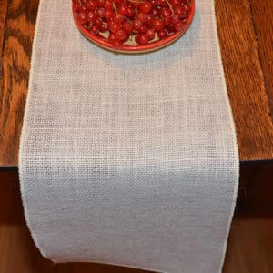 A white burlap table runner on a wooden table. There is a dish with red cherries on top of the burlap runner.