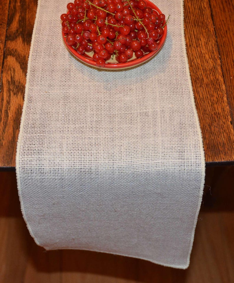 A white burlap table runner on a wooden table. There is a dish with red cherries on top of the burlap runner.