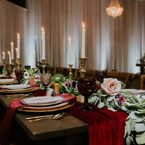A long wooden table set for a formal dinner with red velvet table runners, gold chargers, white plates, and gold flatware. The centerpiece features a garland of greenery, pink roses, and burgundy candlesticks with white candles.