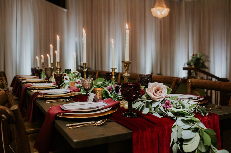 A long wooden table set for a formal dinner with red velvet table runners, gold chargers, white plates, and gold flatware. The centerpiece features a garland of greenery, pink roses, and burgundy candlesticks with white candles.