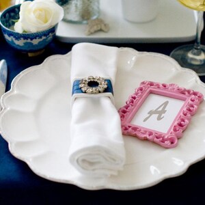 A white dinner plate with a white napkin tied with a blue ribbon and a rhinestone buckle. A pink picture frame with the letter 'A' inside sits on the plate. A silver knife and fork are on the table.