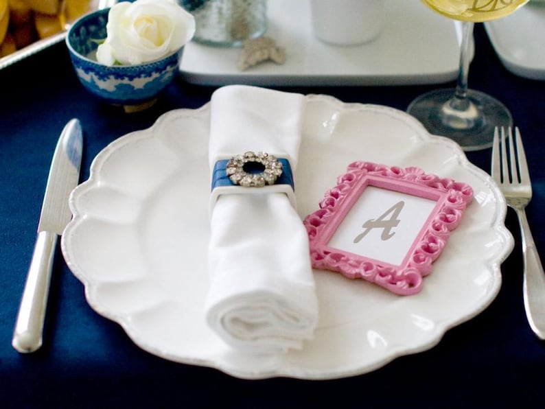 A white dinner plate with a white napkin tied with a blue ribbon and a rhinestone buckle. A pink picture frame with the letter 'A' inside sits on the plate. A silver knife and fork are on the table.