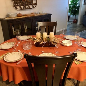 May include: A round orange tablecloth with a centrepiece of three white candles, autumn foliage, and a wooden box. The table is set with white plates, cutlery, and wine glasses. There are two chairs at the table, one of which is in focus.