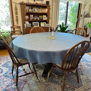 Peut inclure: Une table à manger ronde recouverte d'une nappe bleue, entourée de chaises en bois avec des assises rembourrées. La table est ornée de bougeoirs en laiton. La pièce comprend un tapis à motifs et un buffet en bois.