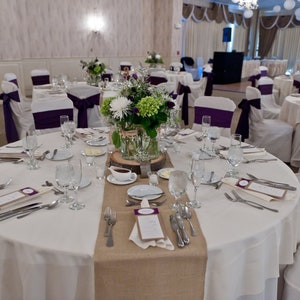 A round table with a white tablecloth and a burlap runner. The table is set with white plates, silverware, and glasses. There is a centerpiece of white and green flowers in a glass vase. The chairs are white with purple sashes.
