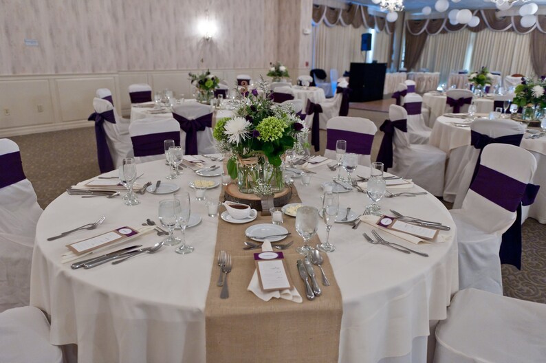 A round table with a white tablecloth and a burlap runner. The table is set with white plates, silverware, and glasses. There is a centerpiece of white and green flowers in a glass vase. The chairs are white with purple sashes.