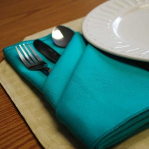 A turquoise napkin folded into a pocket shape with a fork and a spoon inside. The napkin is on a beige placemat.