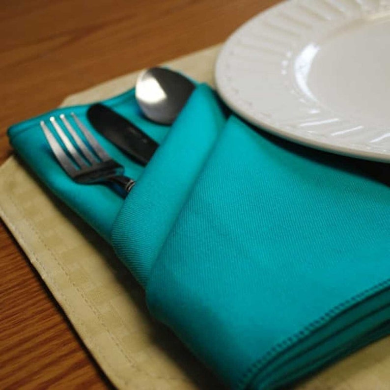 A turquoise napkin folded into a pocket shape with a fork and a spoon inside. The napkin is on a beige placemat.