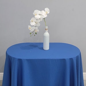 May include: A white painted bottle with a twine-wrapped neck holds a stem of white orchid flowers. The bottle sits on a round, royal blue tablecloth. The background is a plain grey wall.