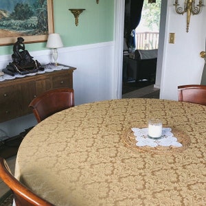 May include: A round dining table with a gold patterned tablecloth. A white candle sits on a woven placemat in the center of the table.