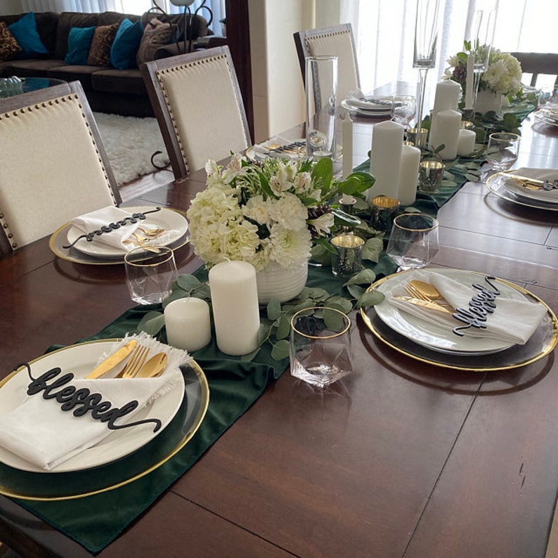 May include: A table setting with a green tablecloth and gold chargers. Each place setting includes a white napkin, gold silverware, a glass, and a black wooden sign that says "blessed".