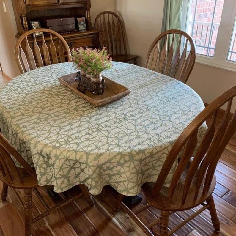 A dining table with a green and white Shibori oval tablecloth. The table is surrounded by wooden chairs. A wooden tray with a decorative vase of flowers sits in the center of the table. A wooden hutch is in the background.