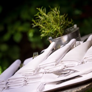 A stack of white linen napkins folded neatly on a table with silverware and a potted plant in the background.
