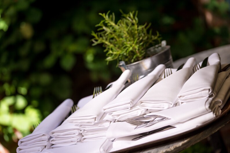 A stack of white linen napkins folded neatly on a table with silverware and a potted plant in the background.