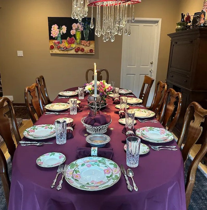 A formal dining table setting with a purple tablecloth, white plates with floral designs, clear glasses, and silver cutlery. A centerpiece with a candle and flowers is on the table.