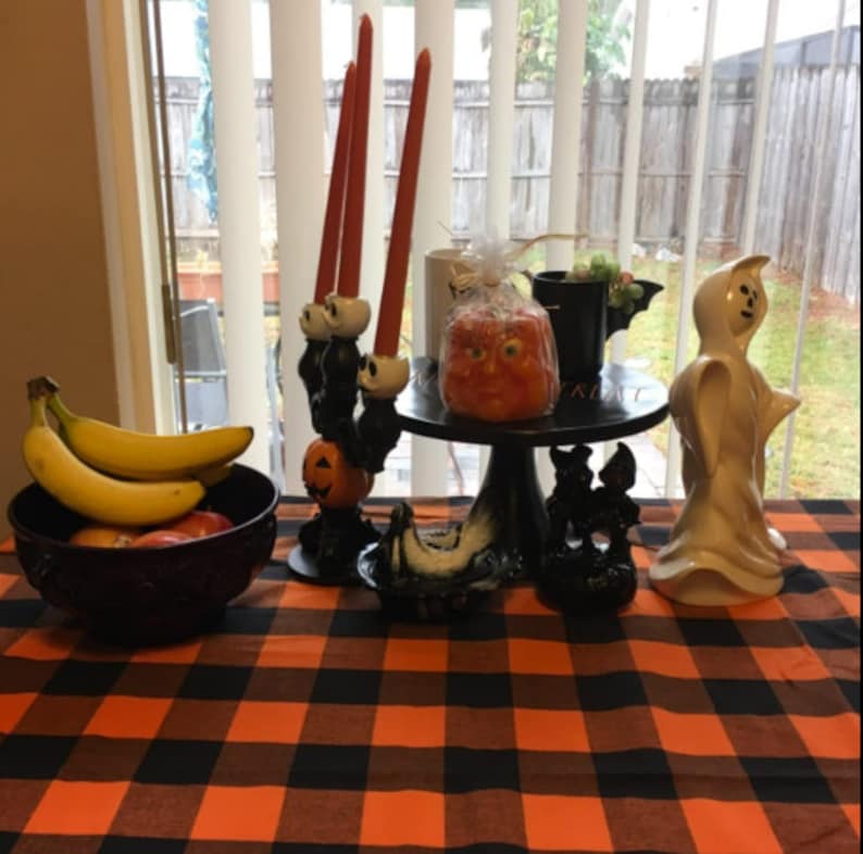 A black and orange checkered tablecloth, also called a plaid tablecloth, with Halloween decorations, including a ghost figurine, a black candle holder with orange candles, a black cake stand with an orange pumpkin-shaped candle, and a bowl of bananas.