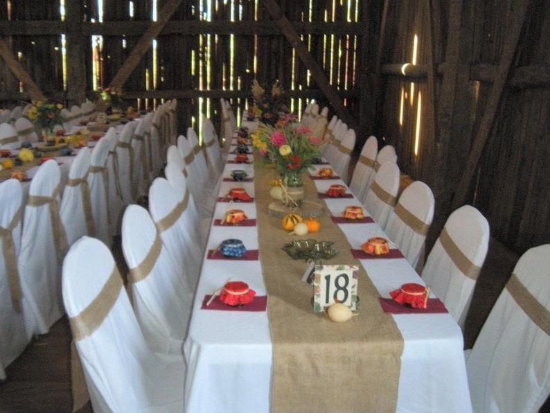 A long table with white linens and burlap runners is set for a wedding reception. The table is decorated with flowers and has place cards with the number 18 on them.