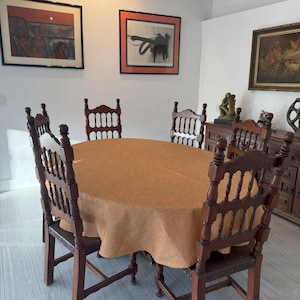May include: A dining room set featuring a round table covered with a tan tablecloth and six wooden chairs. The chairs have a dark brown finish and ornate backs. Artwork hangs on the white walls.