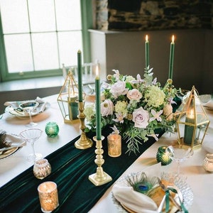 A green velvet table runner with gold candle holders and green candles. A centerpiece of white and green flowers sits on the runner. The table is set with white plates, gold chargers, and clear glasses.