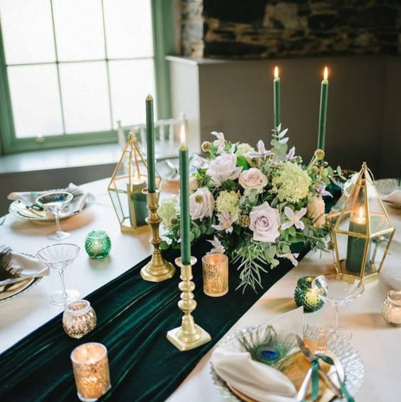 A green velvet table runner with gold candle holders and green candles. A centerpiece of white and green flowers sits on the runner. The table is set with white plates, gold chargers, and clear glasses.