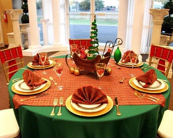 May include: A green tablecloth with a red and gold plaid pattern is set for a holiday dinner. The table is decorated with a miniature Christmas tree in a sleigh, gold ornaments, and red and gold napkins.