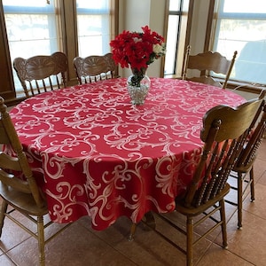 May include: A red round tablecloth with a white damask pattern. The tablecloth is on a round table with six chairs. There are red flowers in a vase in the center of the table.
