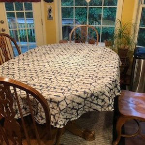 A oval dining table covered with a white and navy blue patterned Shibori tablecloth. The table is surrounded by wooden chairs. A potted plant sits near a window, and the room has a warm, inviting atmosphere.