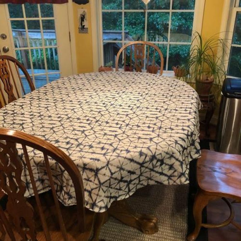 A oval dining table covered with a white and navy blue patterned Shibori tablecloth. The table is surrounded by wooden chairs. A potted plant sits near a window, and the room has a warm, inviting atmosphere.