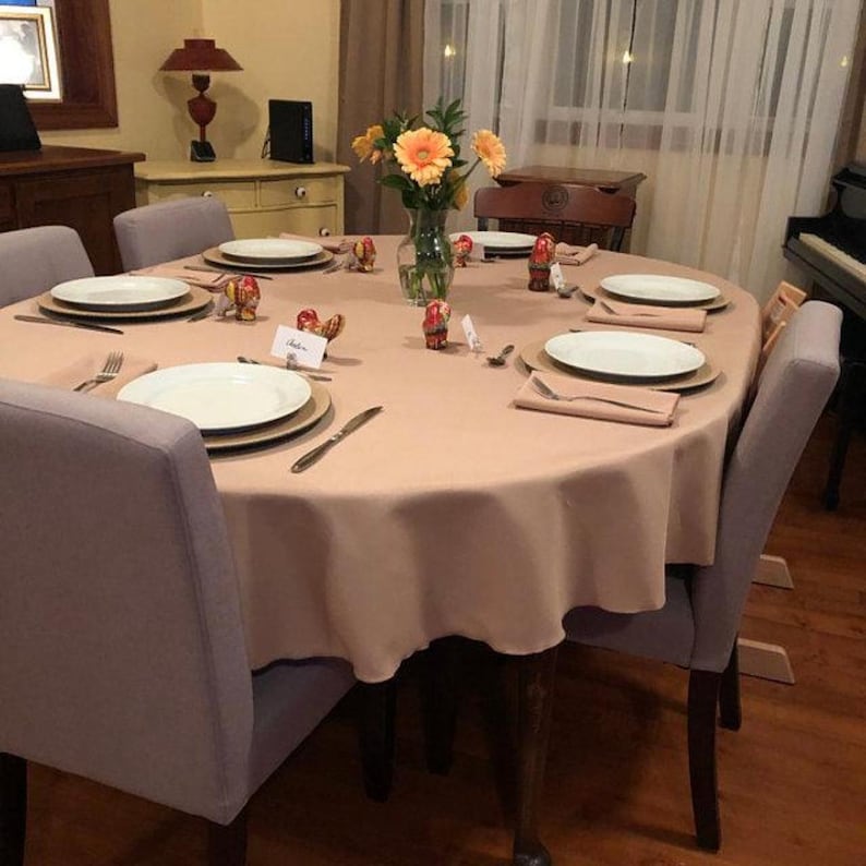 A beige oval tablecloth, folded napkins and placemats under the china in the dining room of a home. Silverware and glasses are also there as the table is ready for an elegant meal.