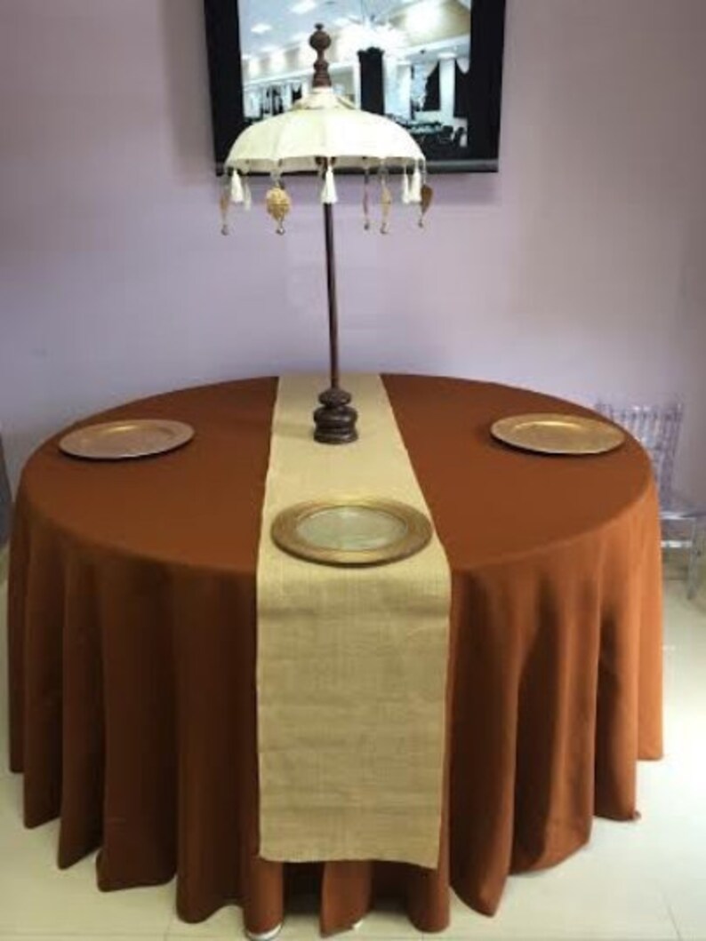 A brown round tablecloth with a burlap table runner and two gold chargers. A white umbrella with gold tassels is in the center of the table.