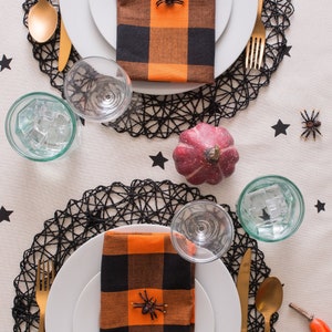 Two Halloween table settings with black and orange checkered napkins, black spider decorations, and gold cutlery on black lace placemats.