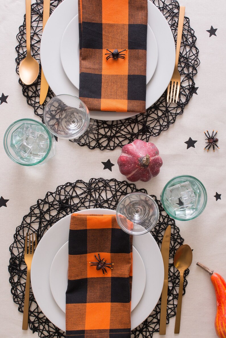 Two Halloween table settings with black and orange checkered napkins, black spider decorations, and gold cutlery on black lace placemats.