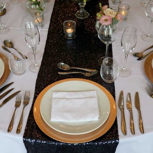 May include: A black sequined table runner with gold rimmed plates and white napkins set on a white tablecloth. The table is decorated with flowers and candles.
