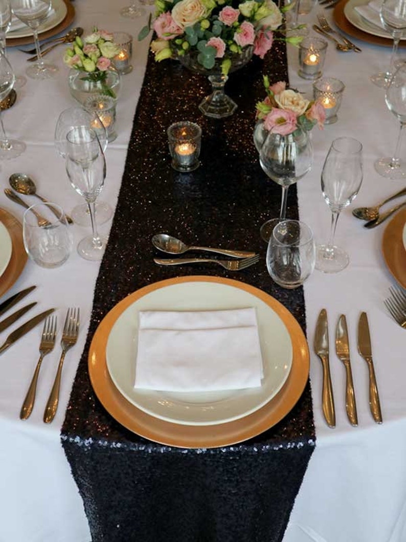 May include: A black sequined table runner with gold rimmed plates and white napkins set on a white tablecloth. The table is decorated with flowers and candles.