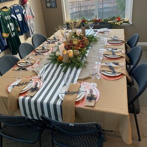 May include: A Thanksgiving table setting with a black and white striped runner and brown placemats. The centerpiece is a green garland with fall foliage and candles. The place settings include white plates, orange chargers, and black cutlery with a 'blessed' and 'grateful' inscription.