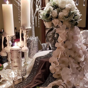 May include: A white floral arrangement with white orchids cascading down from a clear glass vase. The arrangement is on a table with a brown tablecloth and silver accents. There are also white candles and a silver tray with food on the table.