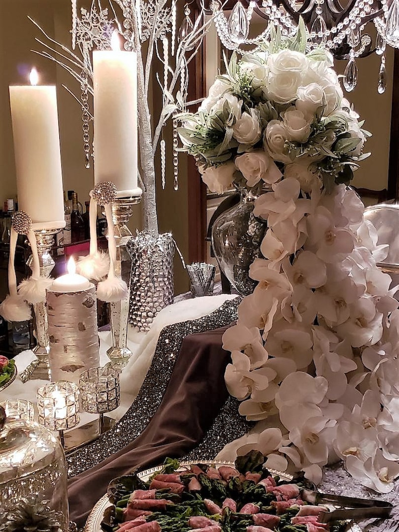 May include: A white floral arrangement with white orchids cascading down from a clear glass vase. The arrangement is on a table with a brown tablecloth and silver accents. There are also white candles and a silver tray with food on the table.
