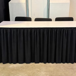 May include: A rectangular table covered with a smooth white top and a pleated black fabric skirt that extends to the floor. Three black chairs are positioned behind the table, with their backrests visible above the white tabletop surface.
