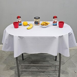 May include: A white oval tablecloth draped over a table with two red cups, two Premier Protein chocolate shake cartons, a yellow lemon, a yellow banana, a jar of On The Border Queso, and a white bowl filled with yellow tortilla chips.