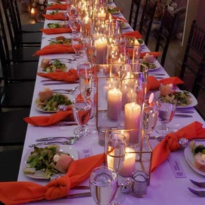 A long table set for a wedding reception with purple velvet table linens, orange napkins, and clear glass candle holders with lit candles. The table is decorated with greenery and flowers.